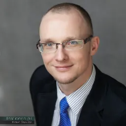 Portret uśmiechniętego mężczyzny w okularach, w garniturze i koszuli w paski z niebieskim krawatem w kropki, na szarym tle.