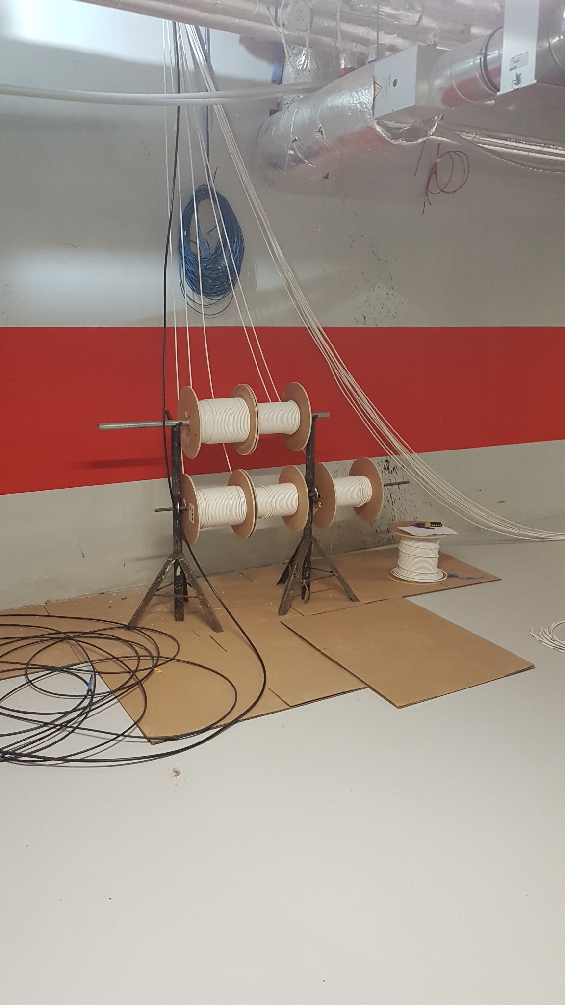 Rozwijanie białych kabli telekomunikacyjnych z drewnianych szpul, zabezpieczonych kartonem, w pomieszczeniu z czerwoną linią na ścianie i widoczną instalacją pod sufitem.