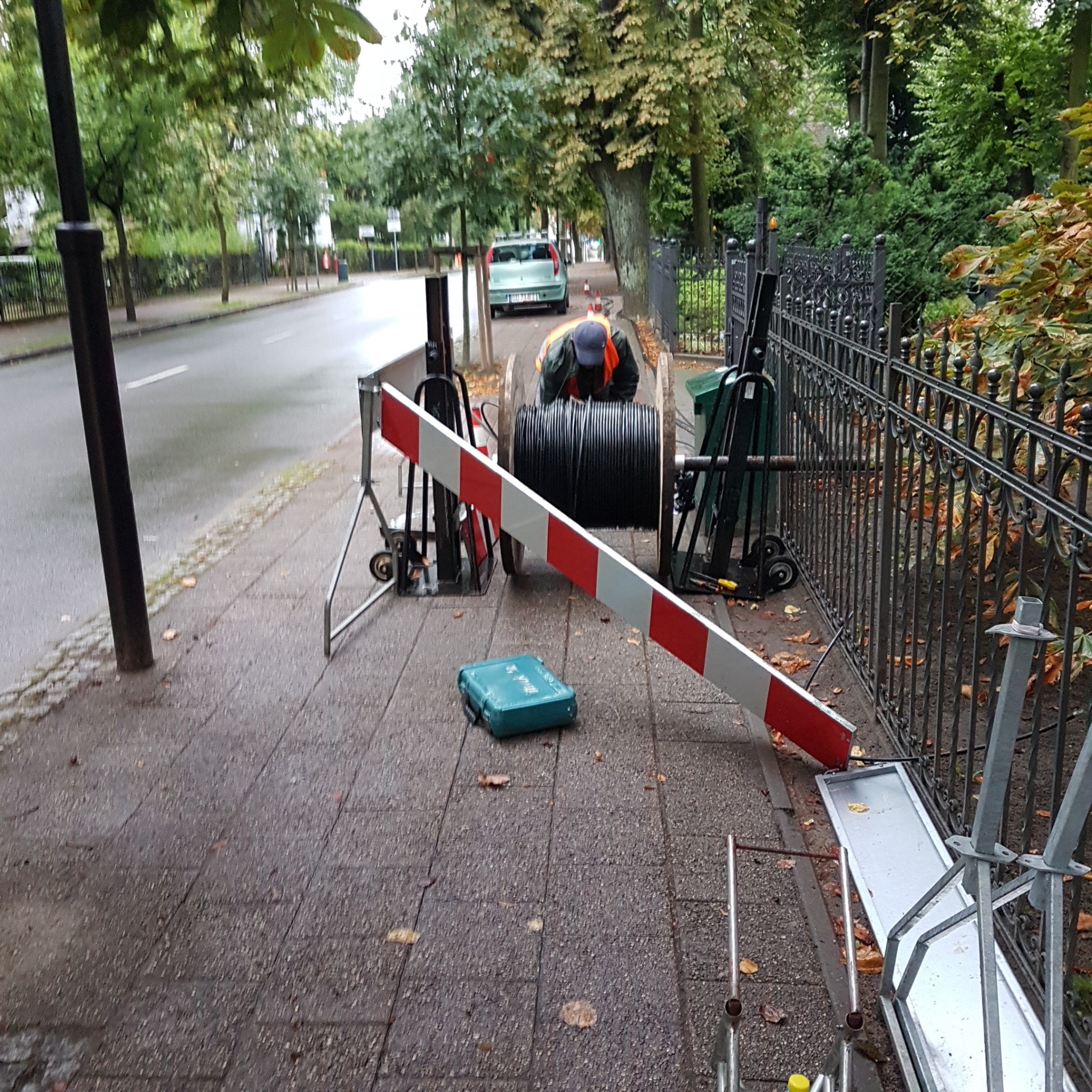 Pracownik w kamizelce odblaskowej pracuje przy dużej szpuli z czarnym kablem na chodniku, obok skrzynka narzędziowa i barierka z czerwono-białymi pasami.