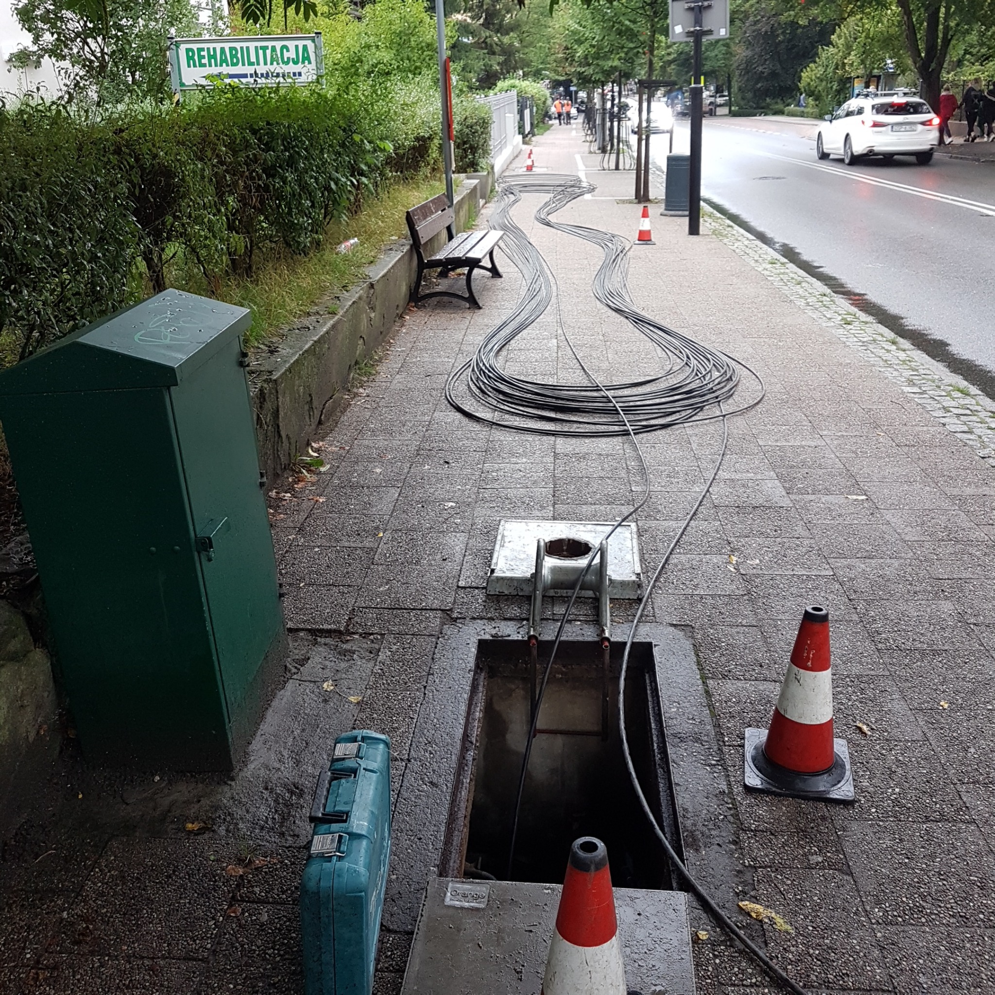 Otwarta studzienka telekomunikacyjna na chodniku z rozłożonymi kablami, skrzynka narzędziowa obok, a w tle znak 'Rehabilitacja'.