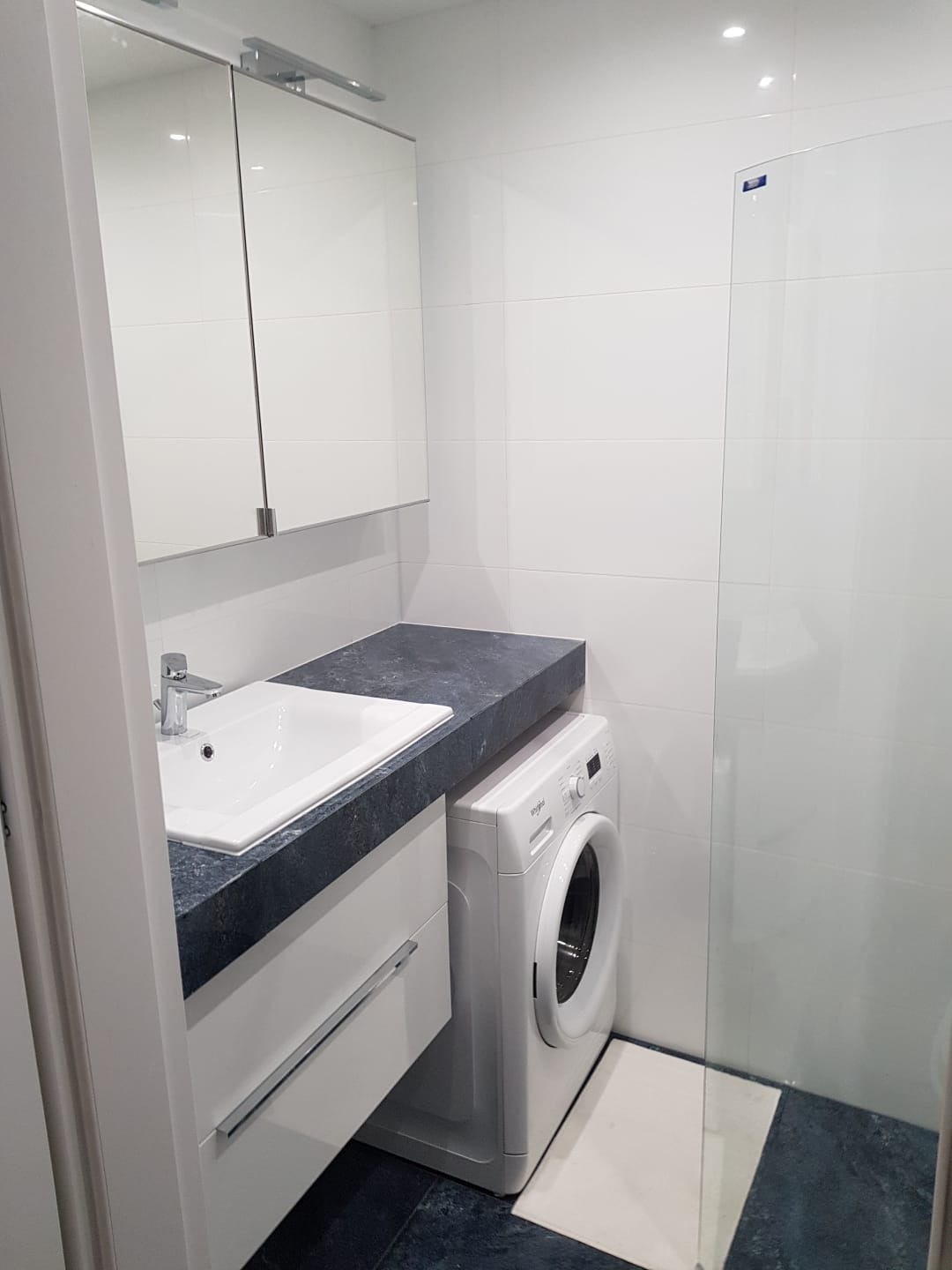 Nowoczesna łazienka z białą umywalką wbudowaną w ciemny blat, białą szafką pod umywalką, pralką obok i kabiną prysznicową w tle, wykończona białymi płytkami.