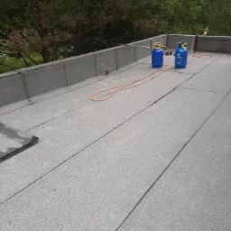 Nowy, płaski dach pokryty papą termozgrzewalną, widoczne butle gazowe i przewód gazowy, tło z zielenią drzew.