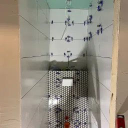 Wnęka prysznicowa w trakcie układania płytek: białe płytki na ścianach i czarno-białe z geometrycznym wzorem na podłodze, niebieskie klipsy poziomujące, widoczne przewody elektryczne i poziomica.