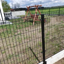 Czarny panelowy płot z betonową podmurówką na tle zielonego trawnika i drewnianej huśtawki ogrodowej, widok z bliska na słupek mocujący.