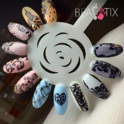 Paleta z wzorami zdobień paznokci w różnych kolorach, z czarnymi ornamentami i złotymi koralikami, prezentowana na białym uchwycie z logo firmy.