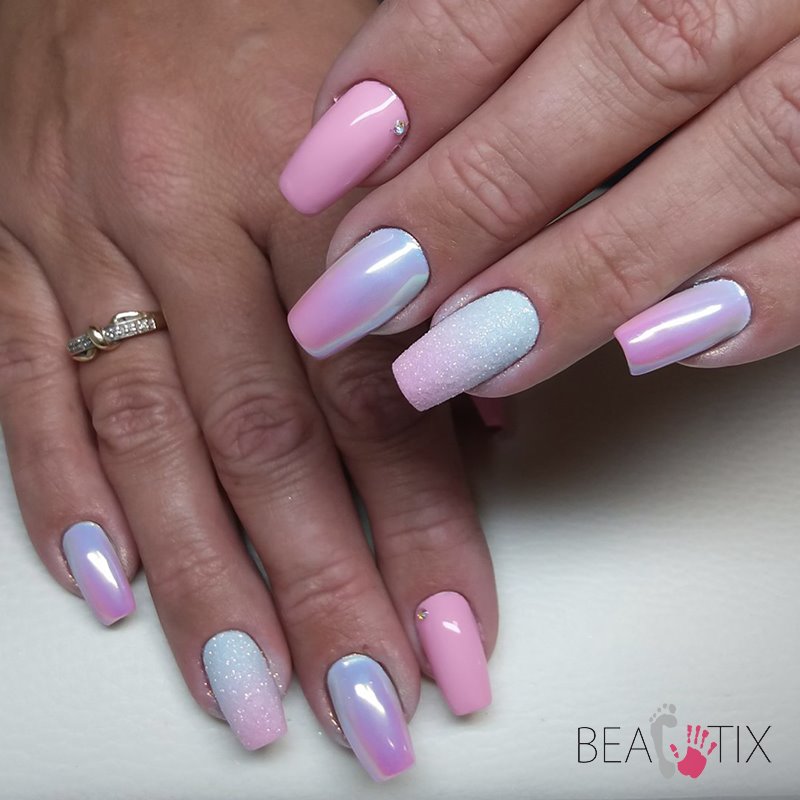 Dłonie z pomalowanymi paznokciami w odcieniach różu i bieli, z efektem opalizującym i brokatowym, jeden paznokieć ozdobiony drobnym kryształkiem, widoczny pierścionek na palcu.