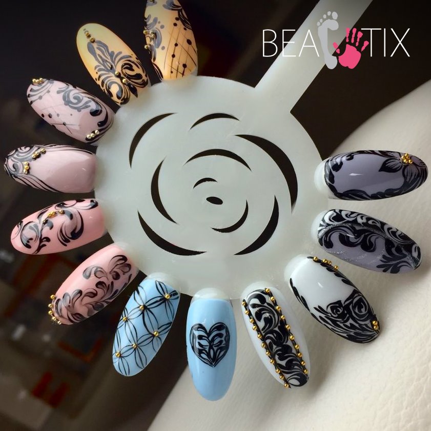 Paleta z wzorami zdobień paznokci w różnych kolorach, z czarnymi ornamentami i złotymi koralikami, prezentowana na białym uchwycie z logo firmy.