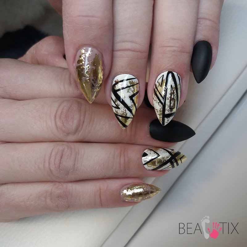 Zbliżenie na dłoń z manicure hybrydowym: migdałki w kolorach złota, bieli i czerni z geometrycznymi wzorami i złotymi płatkami.