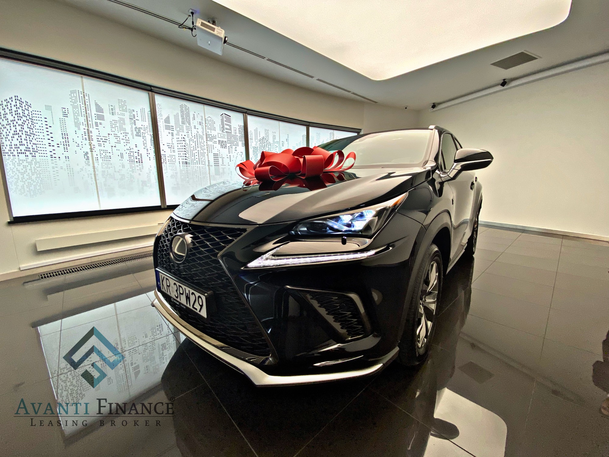 Czarny samochód Lexus NX z czerwoną kokardą na masce, prezentowany w salonie z oknami z motywem miasta i logo firmy Avanti Finance odbitym w podłodze.