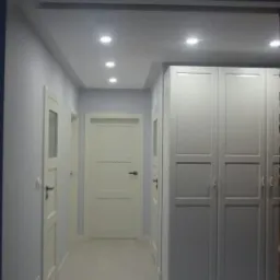 Korytarz po remoncie z białymi drzwiami, szarą szafą w zabudowie i punktowym oświetleniem sufitowym.