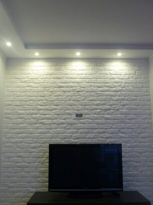 Biała ściana z cegieł dekoracyjnych podświetlona punktowymi lampami sufitowymi, z zamontowanym telewizorem na czarnej szafce RTV.