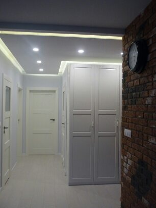 Korytarz z białymi drzwiami, szafą w zabudowie i ceglaną ścianą z zegarem, oświetlenie sufitowe punktowe i ledowe