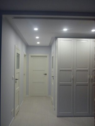 Korytarz po remoncie z białymi drzwiami, szarą szafą w zabudowie i punktowym oświetleniem sufitowym.