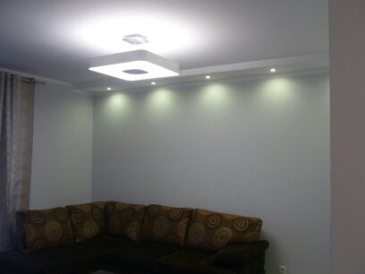 Jasne wnętrze salonu z szarą ścianą, oświetleniem punktowym LED na suficie i geometryczną lampą sufitową, widoczny narożnik sofy z brązowymi poduszkami w tle.