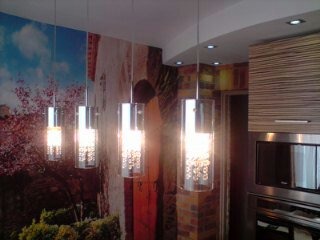 Nowoczesna kuchnia z fototapetą przedstawiającą krajobraz toskański, oświetlona czterema wiszącymi lampami z chromowanymi elementami i kryształkami, zabudowa kuchenna z frontami w odcieniach brązu...