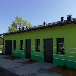 Budynek z zieloną elewacją i ciemnymi drzwiami, ozdobiony poziomymi pasami, z czarnym dachem i kominami, otoczony niewielkimi drzewkami i chodnikiem.