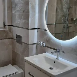 Nowoczesna łazienka z beżowymi płytkami imitującymi marmur, okrągłym lustrem z podświetleniem LED, białą szafką pod umywalką z dwoma szufladami i wiszącą toaletą.