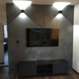 Ściana z efektem dekoracyjnym w odcieniach szarości, oświetlona dwoma czarnymi lampami ściennymi, podwieszony telewizor, ciemna szafka RTV na drewnianej podłodze, widok na pokój przez otwarte drzwi.