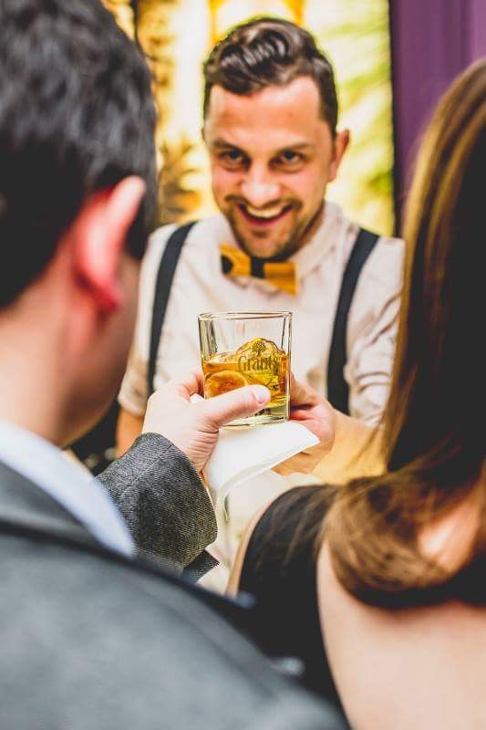 Uśmiechnięty barman w muszce podaje drink z logo Grant's na serwetce, widok zza ramienia klienta.