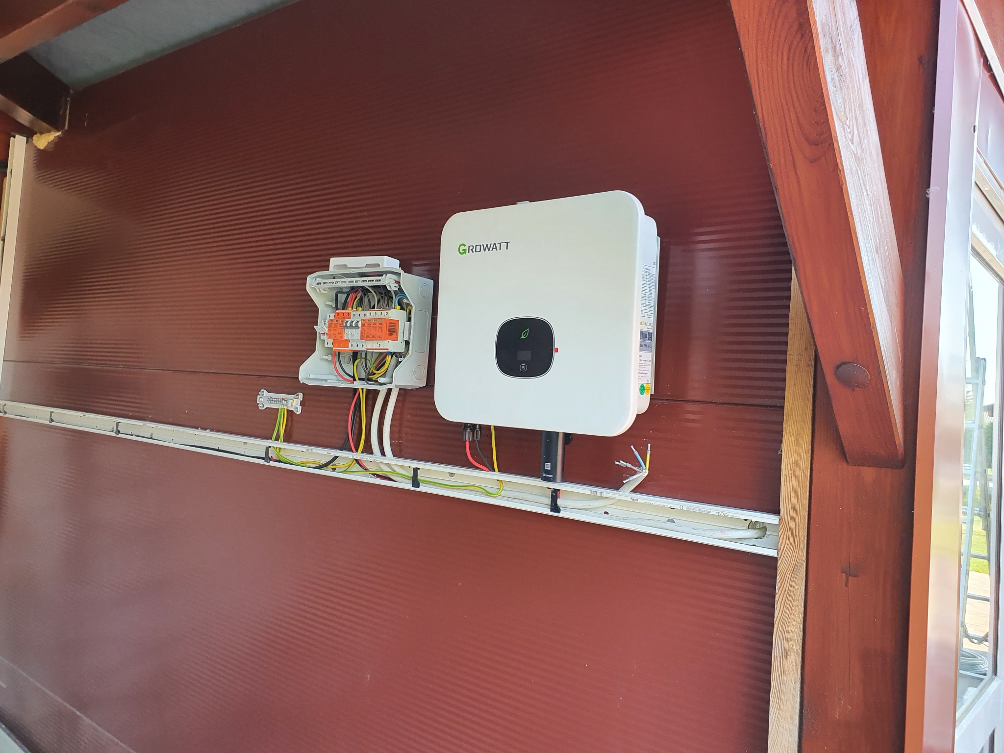 Biały falownik Growatt zamontowany na brązowej ścianie z widoczną skrzynką elektryczną i listwą kablową, instalacja w trakcie podłączania.