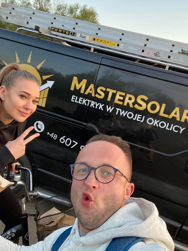 Selfie dwóch osób na tle czarnego samochodu firmy elektrycznej Master Solar z numerem telefonu i drabina na dachu.