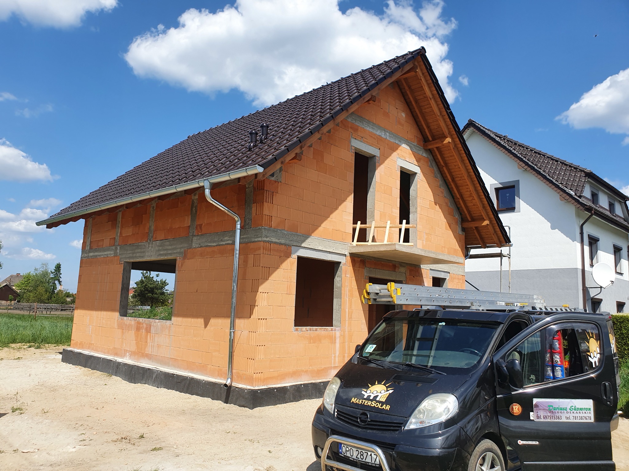 Dom w trakcie budowy z brązowym dachem, widoczne otwory okienne i drzwiowe, obok zaparkowany czarny van z logo firmy budowlanej oraz drabina na dachu, błękitne niebo z chmurami w tle.