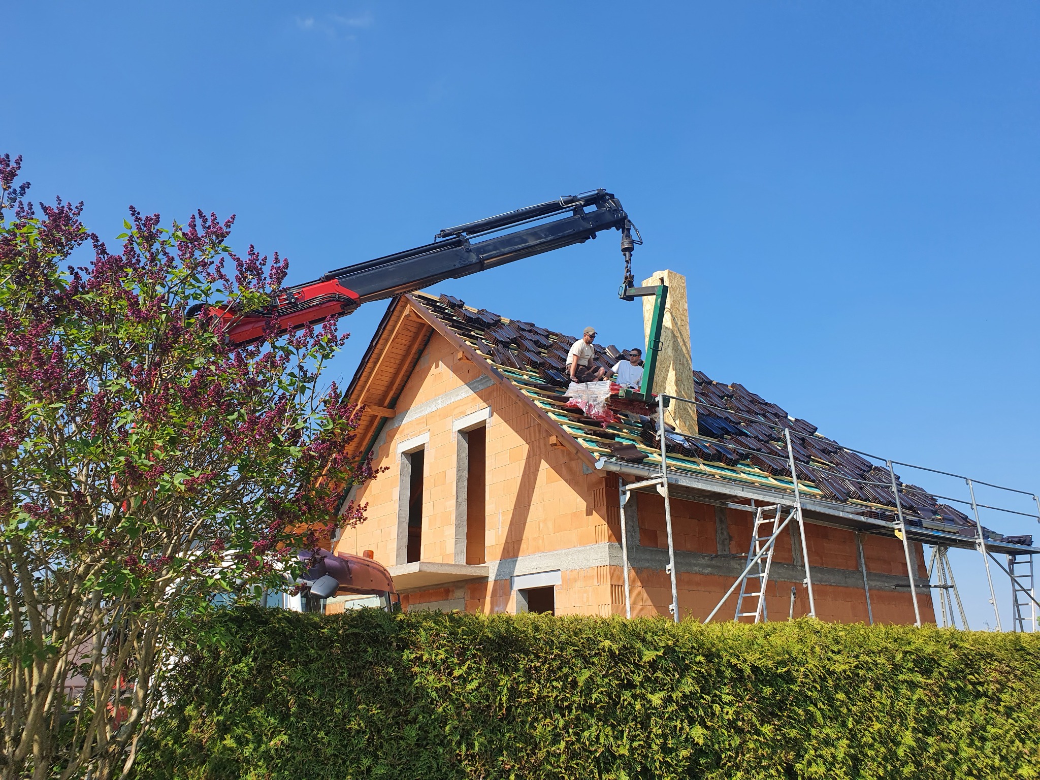 Dwóch dekarzy na szczycie ceglanego domu w trakcie układania ciemnobrązowej dachówki, wspieranych przez dźwig z czerwoną podstawą, z widocznym kominem i rusztowaniem, na tle błękitnego nieba...