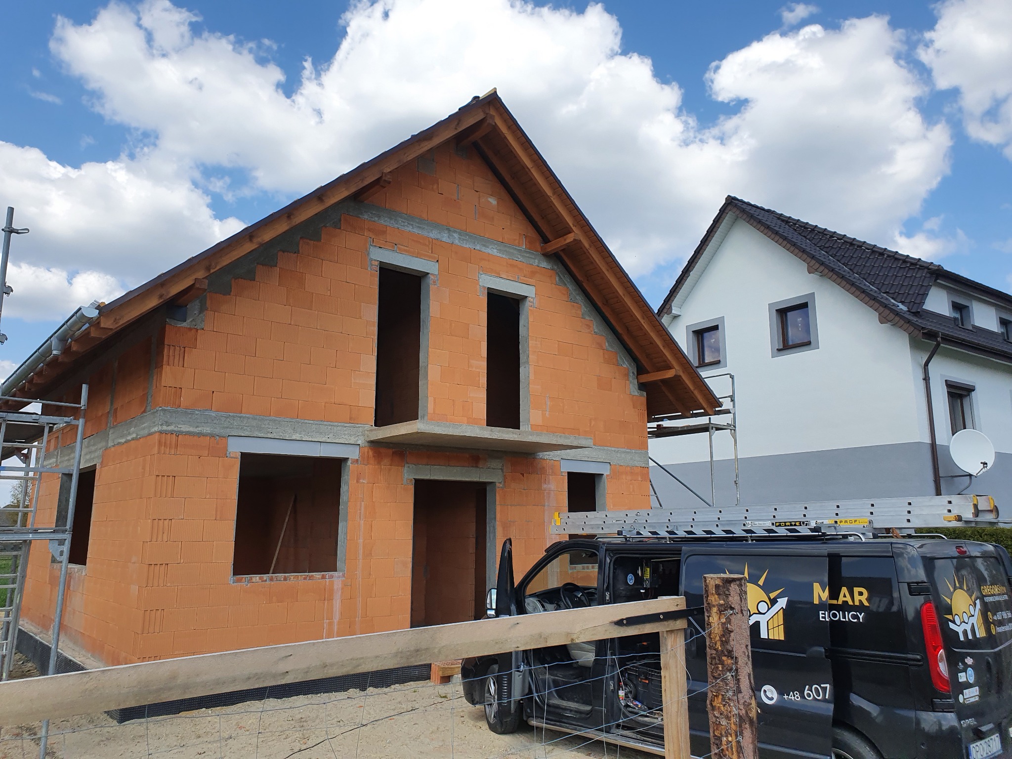 Surowy stan domu z czerwonej cegły w trakcie budowy, widoczny szkielet dachu, okna i drzwi bez osadzenia, rusztowanie z lewej strony, czarny samochód dostawczy z logo firmy w tle, niebo z chmurami.