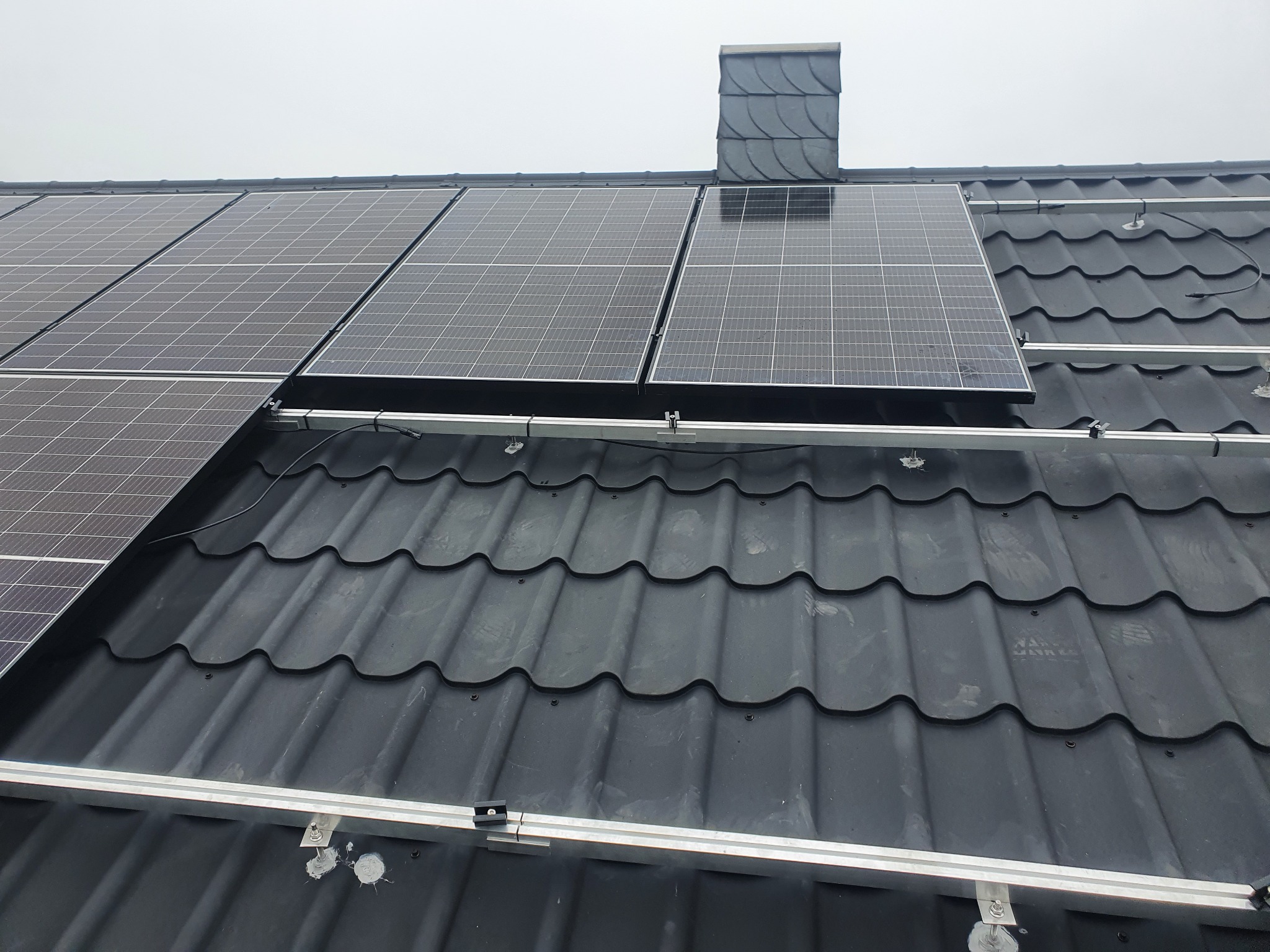 Montaż paneli fotowoltaicznych na dachu z czarnej blachodachówki, widoczne aluminiowe szyny montażowe i komin obłożony łupkiem.