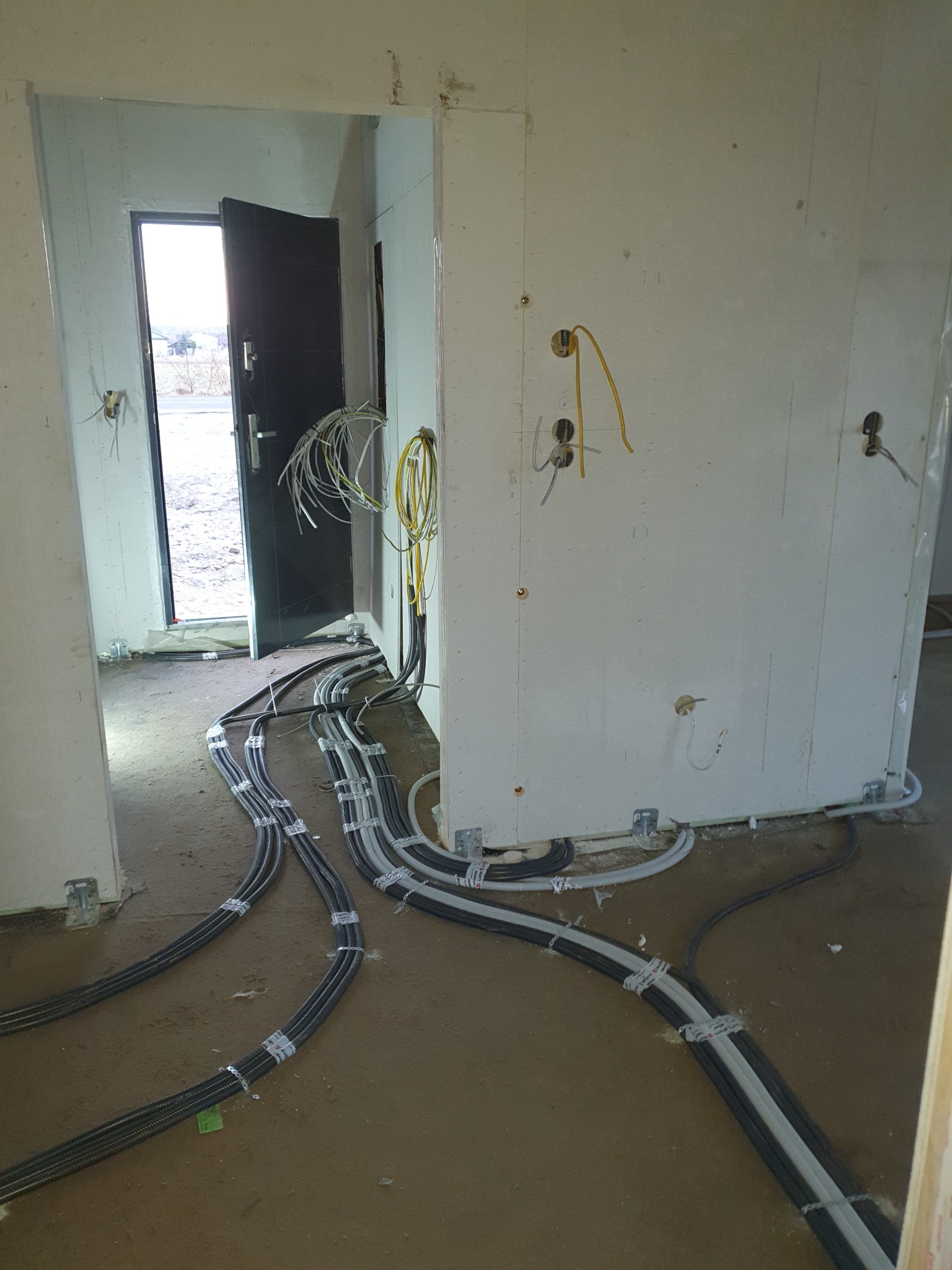 Wnętrze w stanie surowym z widocznymi, licznymi przewodami elektrycznymi ułożonymi na podłodze i wychodzącymi ze ściany, zabezpieczone peszlami i uchwytami mocującymi, z otwartymi drzwiami...