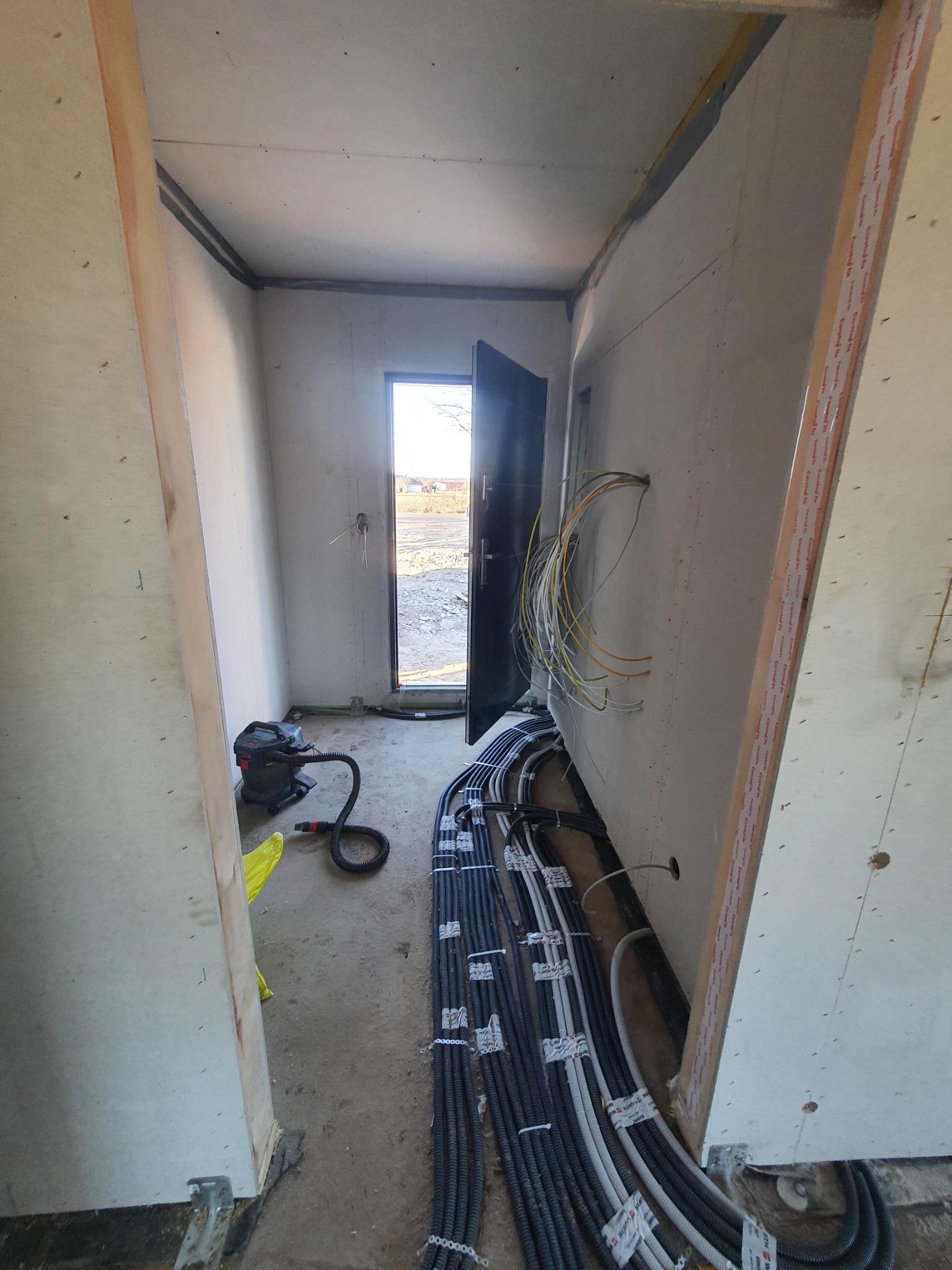 Wnętrze w stanie surowym z widocznymi przewodami elektrycznymi ułożonymi w korytach wzdłuż ściany, częściowo wystającymi ze ściany, odkurzacz przemysłowy na podłodze, otwarte drzwi wejściowe...