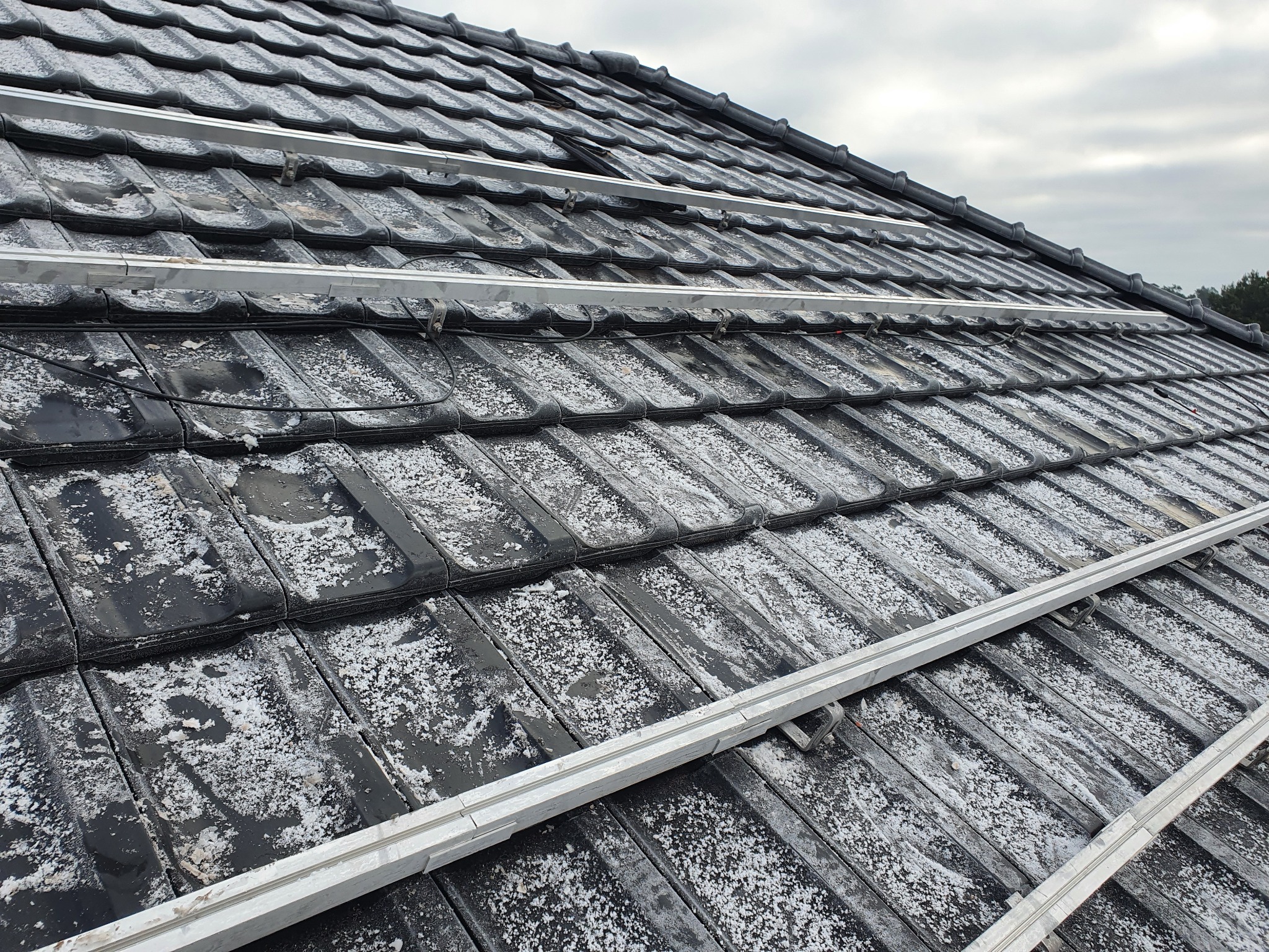 Częściowo zamontowany system szyn montażowych na czarnych dachówkach ceramicznych, przygotowany do instalacji paneli fotowoltaicznych, pokryty białym osadem.