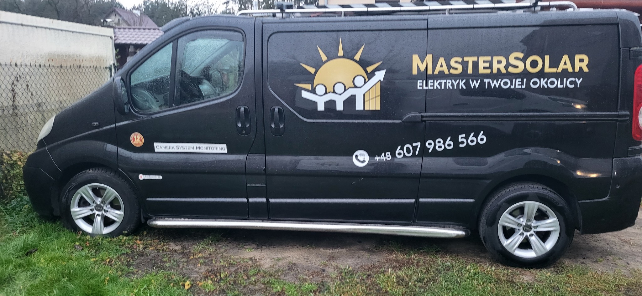 Czarny samochód dostawczy z logo firmy MasterSolar i numerem telefonu, zaparkowany na trawie przed budynkiem.
