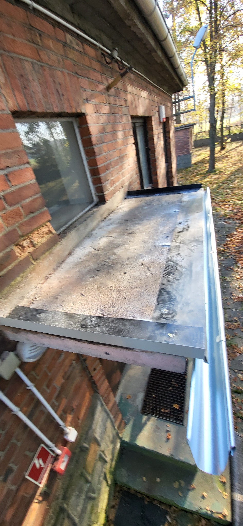 Zewnętrzny widok budynku z cegły z nową, błyszczącą rynną zamontowaną wzdłuż krawędzi dachu, widoczna również instalacja antenowa na ścianie budynku.