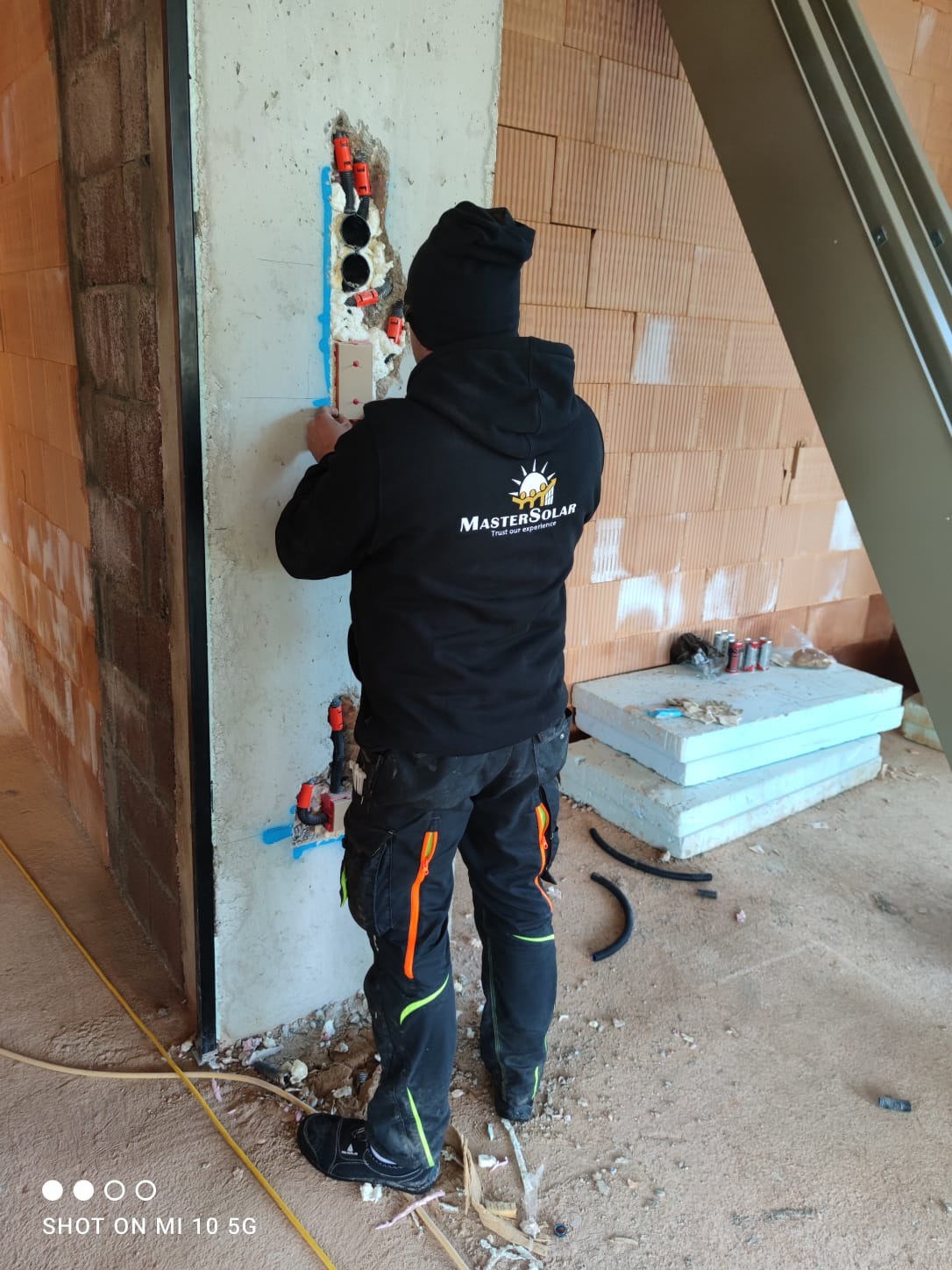 Instalator w czarnej bluzie z logo firmy MASTER SOLAR, montuje rury w ścianie z pustaków, używając pianki montażowej, widoczne przewody i narzędzia, surowy stan wykończenia wnętrza.