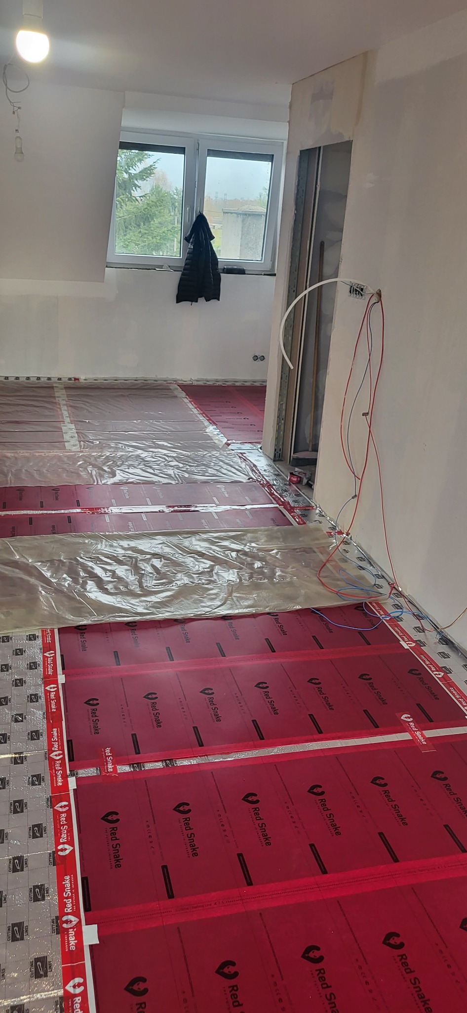 Instalacja folii grzewczej Red Snake na podłodze w trakcie remontu, widoczne przewody elektryczne wychodzące ze ściany, częściowo przykryte folią ochronną.