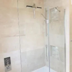 Nowoczesna łazienka z beżowymi płytkami, szklanym parawanem nawannowym z chromowanymi zawiasami, deszczownicą i wnęką wyłożoną mozaiką.