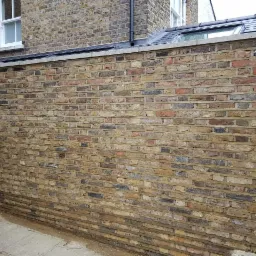 Świeżo wymurowana ściana z żółtej cegły z widoczną zaprawą, fragment elewacji budynku z oknem na górze, drewniany słupek oparty o ścianę.