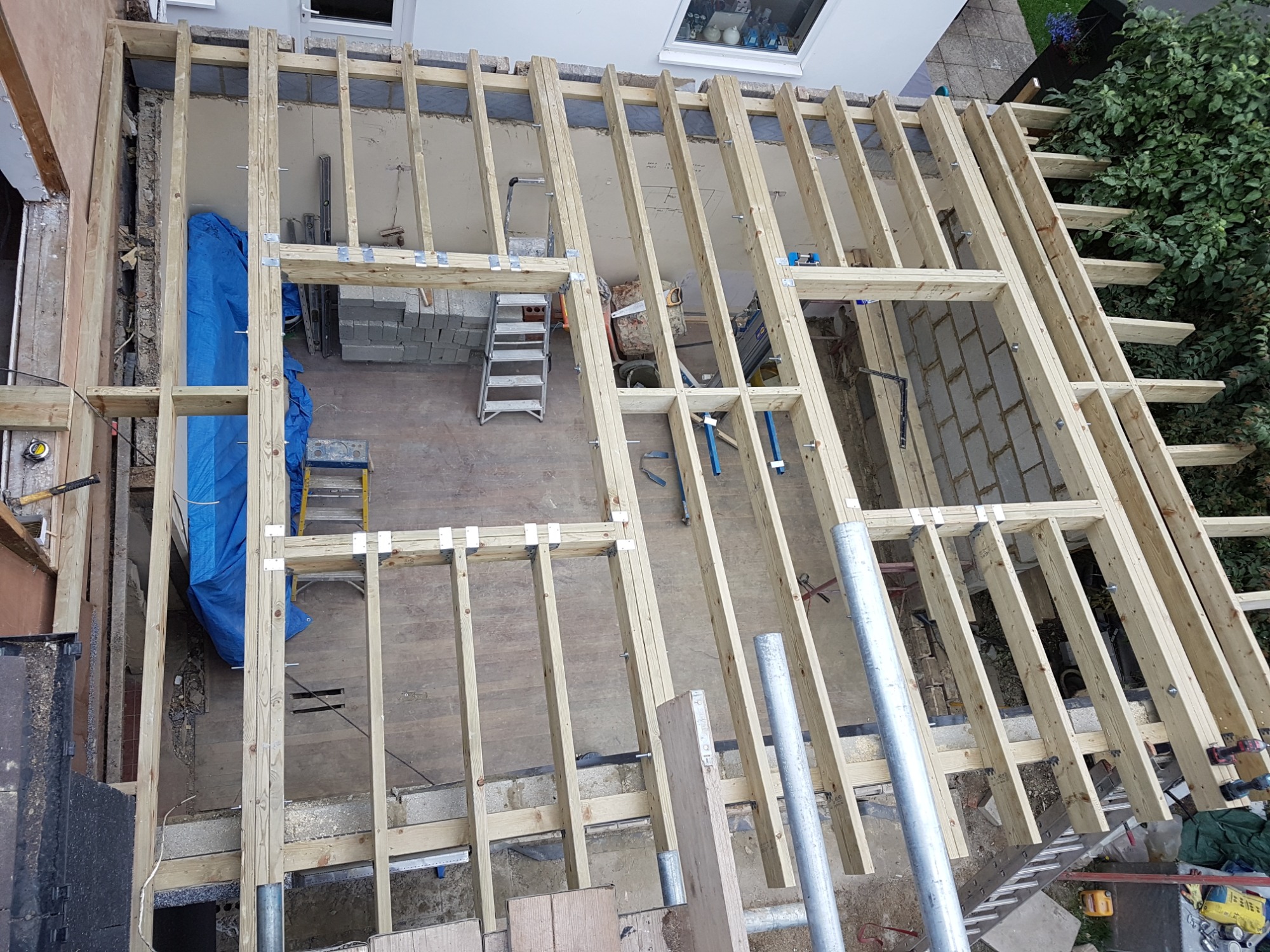 Konstrukcja drewniana dachu w trakcie rozbudowy domu, widok z góry na szkielet z belek, z fragmentami muru i narzędziami budowlanymi w tle.