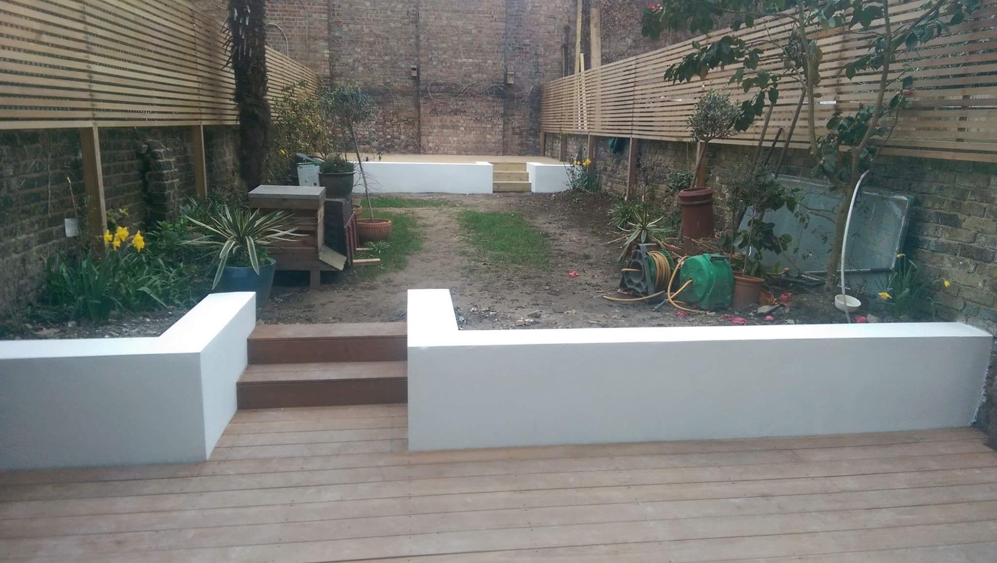 Nowoczesny taras z drewnianą podłogą i białymi, niskimi murkami, prowadzący schodkami do ogrodu z ceglanym murem w tle.