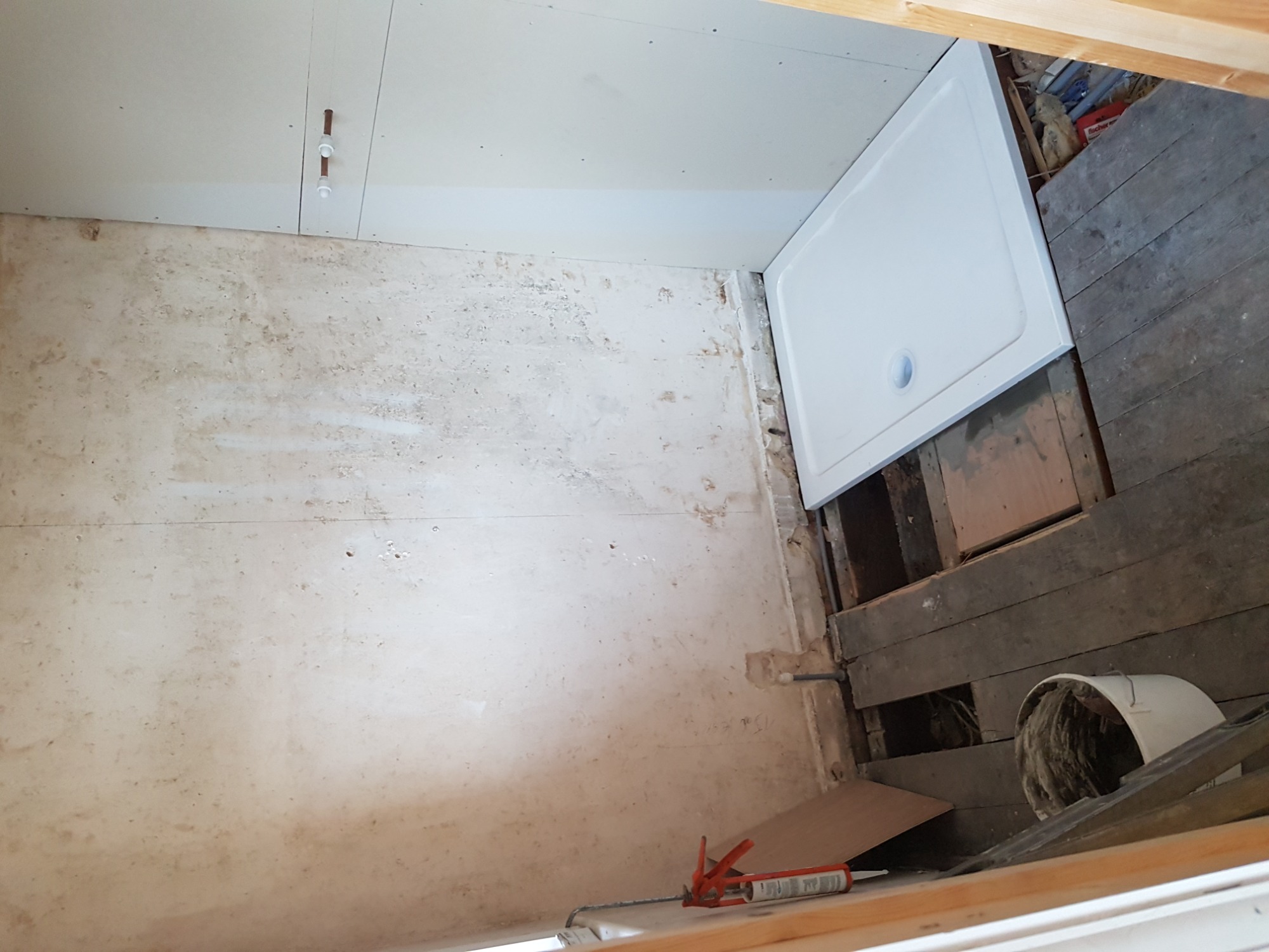 Niedokończona łazienka w trakcie prac: surowa ściana z widocznymi śladami po starych płytkach, częściowo zamontowany brodzik, odsłonięte drewniane elementy konstrukcyjne podłogi, biały sufit...