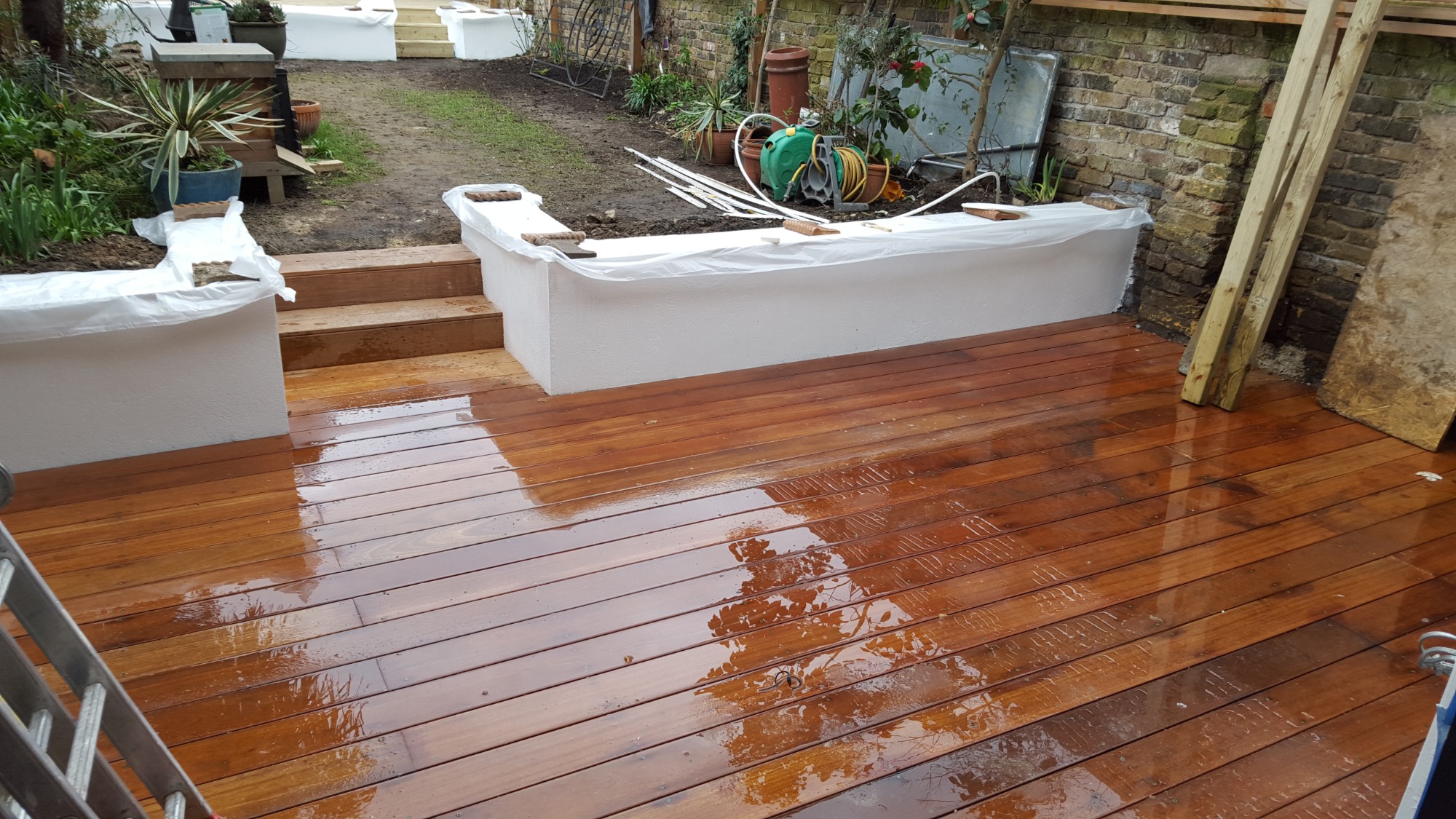 Świeżo położony, mokry drewniany taras z widocznymi śladami po deszczu, otoczony białymi podwyższonymi rabatami kwiatowymi i schodami, w tle ogród z roślinami i ceglanym murem.