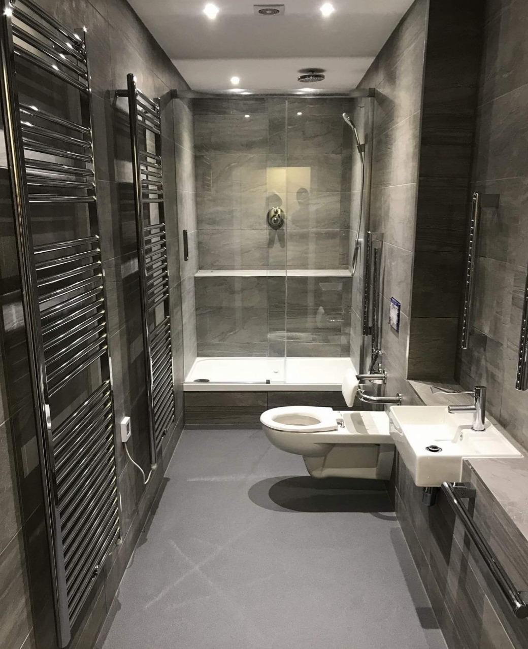 Nowoczesna łazienka z szarymi płytkami imitującymi kamień, kabiną prysznicową typu walk-in, wiszącą toaletą i umywalką, oraz grzejnikami drabinkowymi, przystosowana dla osób niepełnosprawnych...