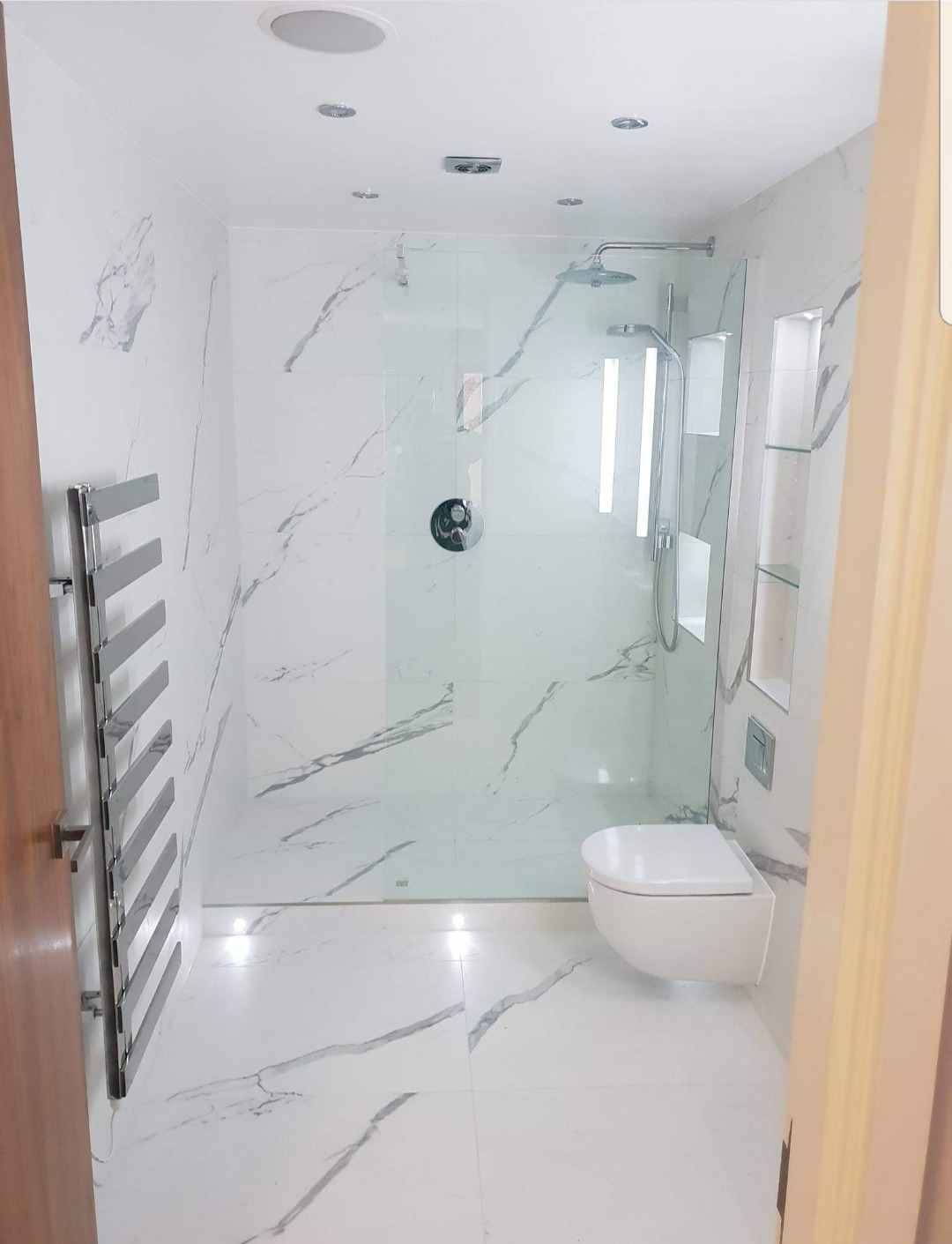 Nowoczesna łazienka z białymi marmurkowymi płytkami, kabiną prysznicową ze szklaną ścianką, podwieszaną toaletą i grzejnikiem drabinkowym, oświetlona punktowymi lampami w suficie i podłodze.