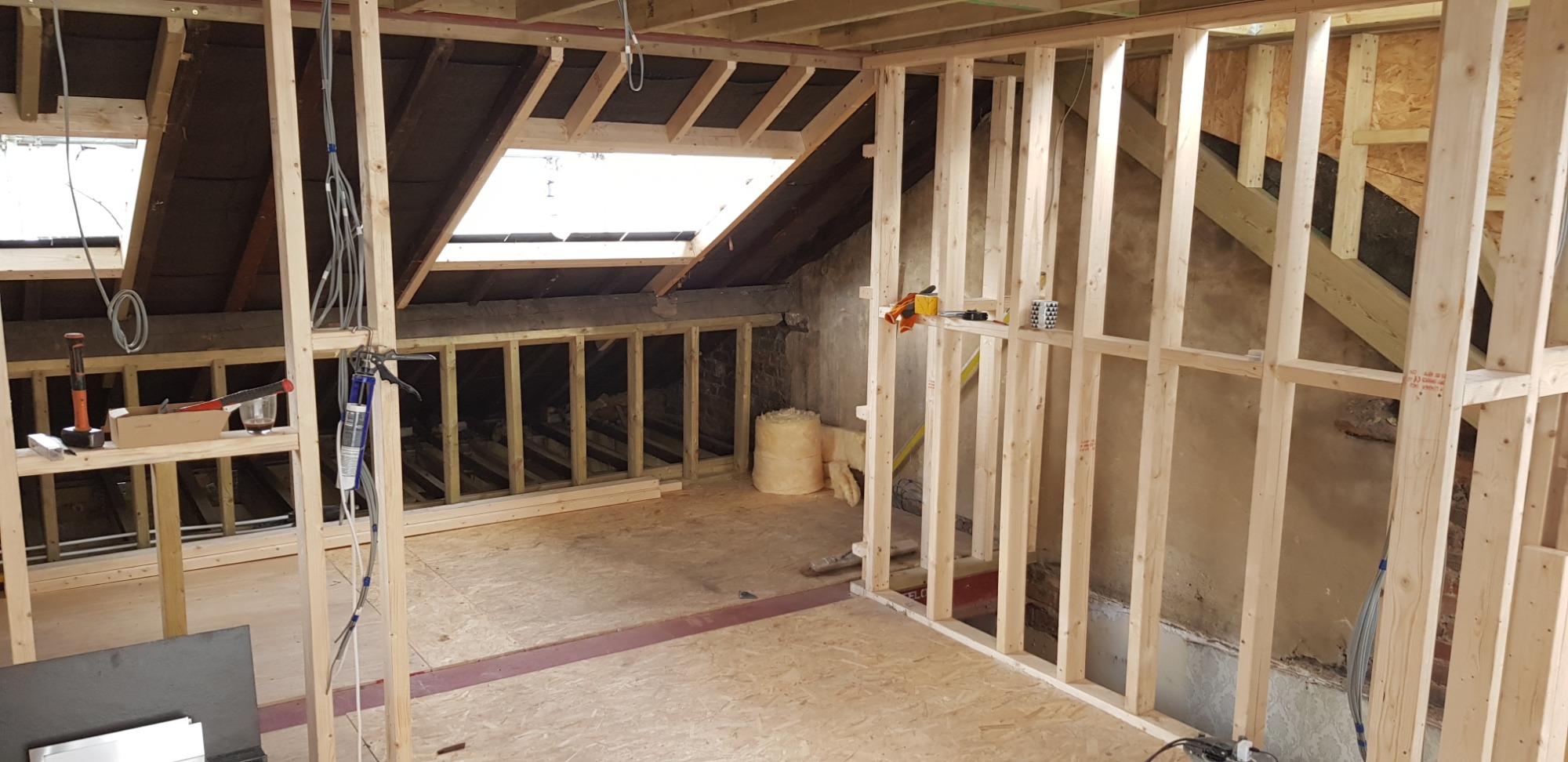 Wnętrze poddasza w trakcie adaptacji: drewniana konstrukcja ścian działowych, okno dachowe, widoczne przewody elektryczne, ocieplenie w rolce i narzędzia pracy.