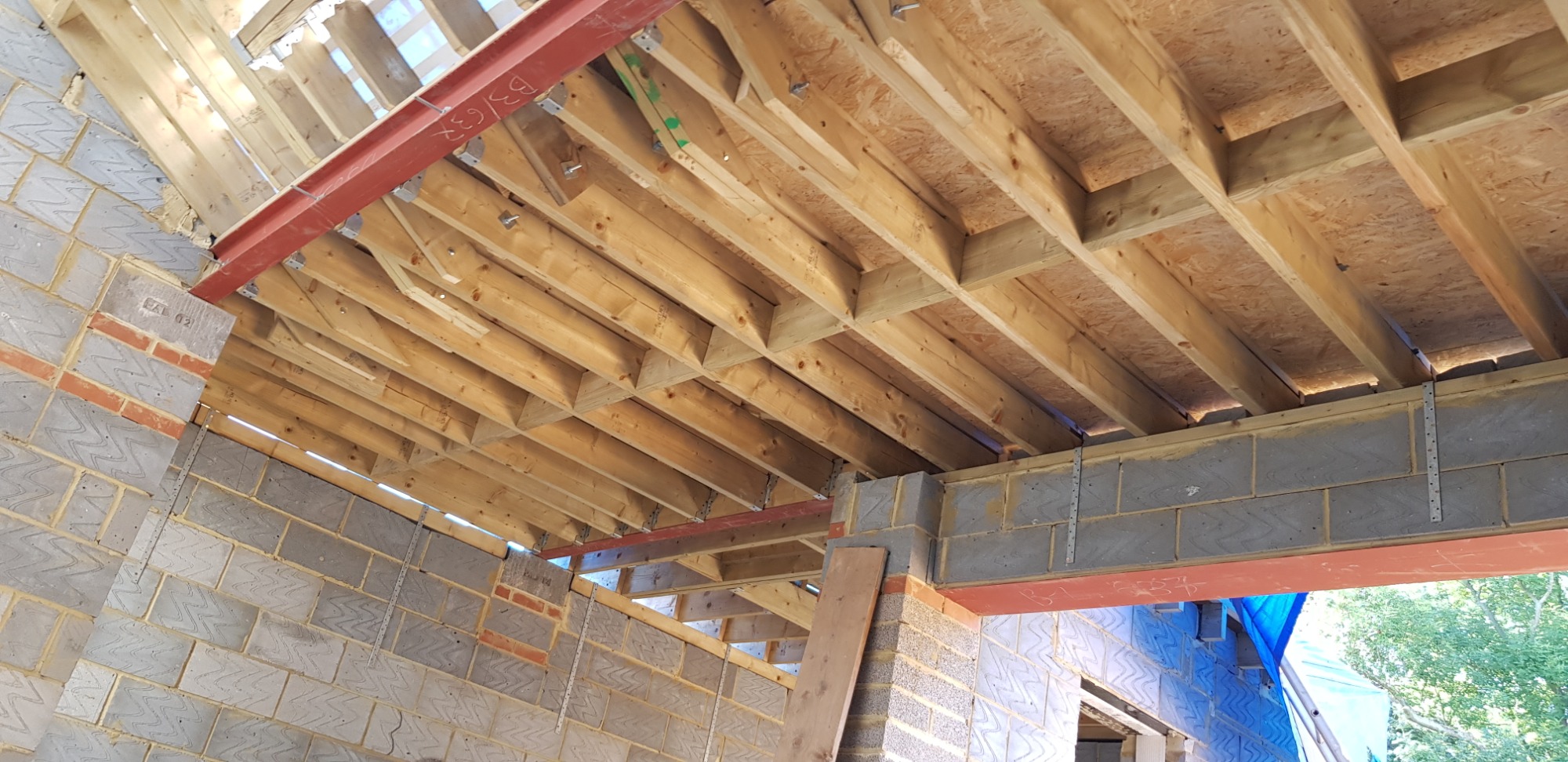 Surowy stan rozbudowy domu: widoczne bloczki betonowe, drewniana konstrukcja stropu z deskowaniem, stalowy profil wzmacniający, mocowania metalowe, częściowo zakryte okno folią ochronną.