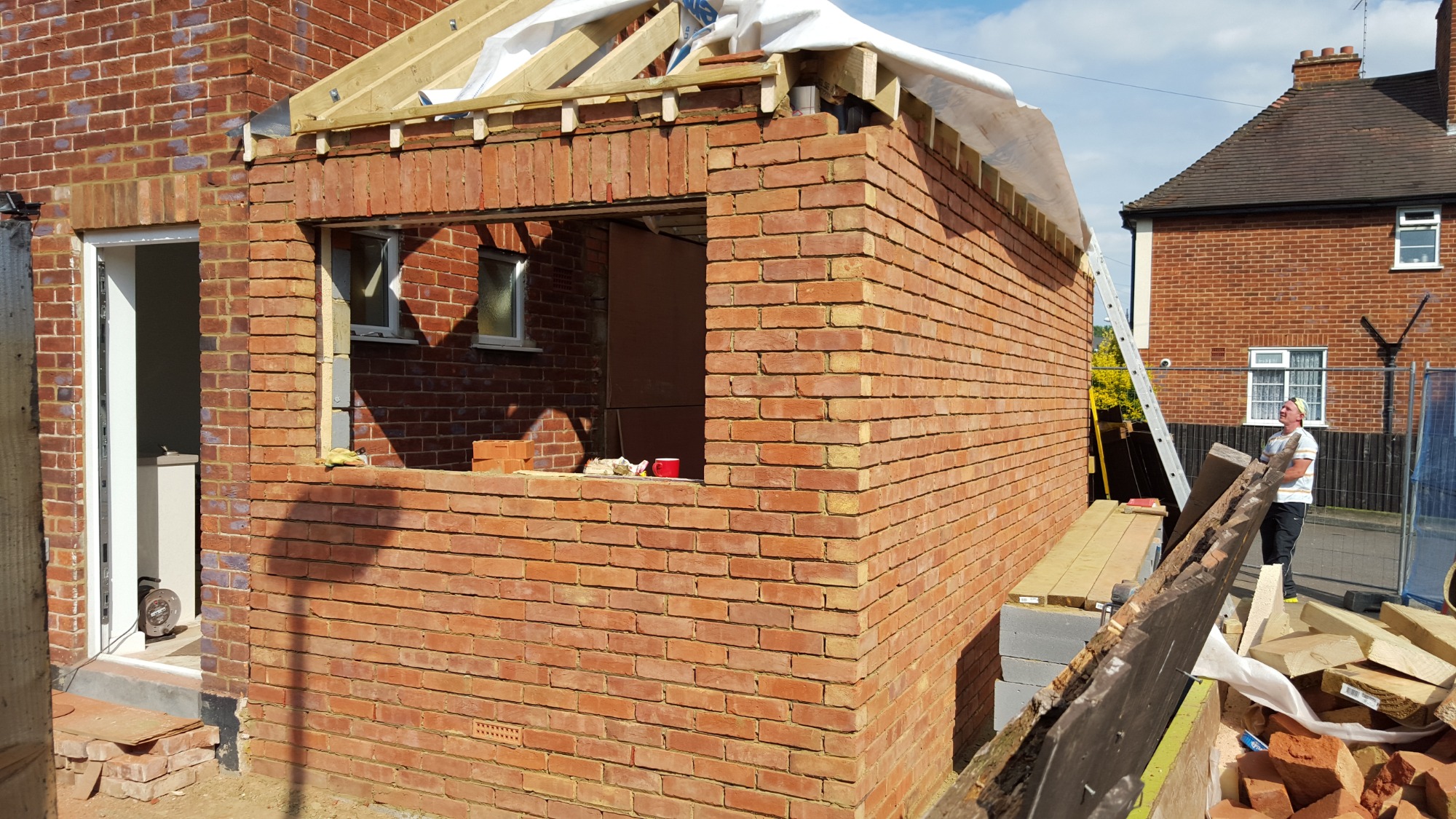 Murowana ściana dobudowy domu z widocznym oknem i drzwiami, częściowo zakryta folią, z pracownikiem na zewnątrz i drabiną opartą o ścianę.
