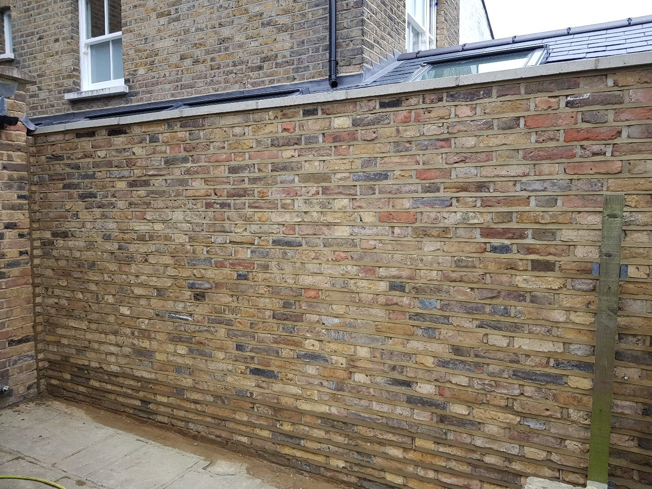 Świeżo wymurowana ściana z żółtej cegły z widoczną zaprawą, fragment elewacji budynku z oknem na górze, drewniany słupek oparty o ścianę.