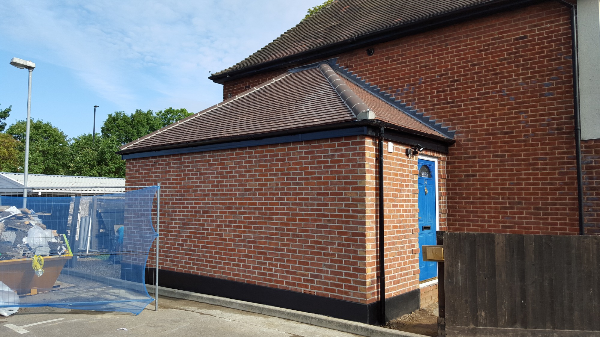 Nowo dobudowana, ceglana część domu z niebieskimi drzwiami wejściowymi, widok z zewnątrz, z widocznym kontenerem na odpady budowlane za siatką.