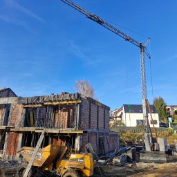 Paweł Wąchała PW usługi remontowo-budowlane - Budowa domu w stanie surowym otwartym, z widocznym żurawiem i wywrotką na tle błękitnego nieba. W tle zabudowa jednorodzinna i drzewa.